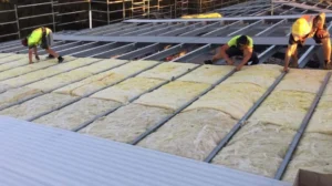 Construction workers install insulation and roofing materials on a commercial building. Wearing high-visibility vests and hard hats, they position thick mineral wool or fiberglass batts between metal roofing supports. This thermal and acoustic insulation is critical for building energy efficiency.